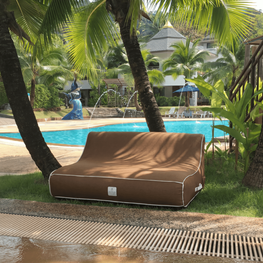 Un canapé gonflable marron est installé sur la pelouse près d'une piscine, sous des palmiers dans un jardin tropical luxueux et ensoleillé.