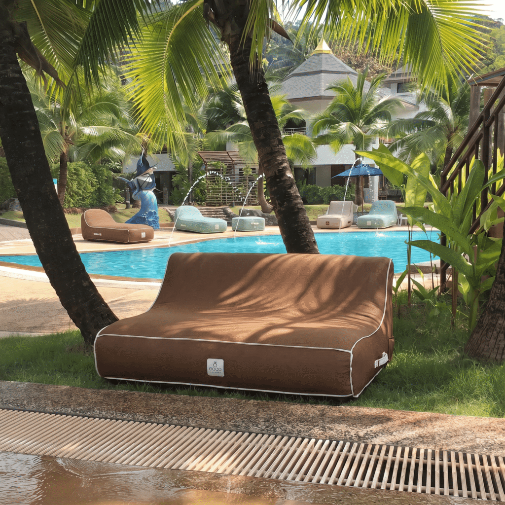 Un transat gonflable marron avec liseré blanc installé sous un palmier au bord d'une piscine tropicale luxuriante avec végétation exotique.