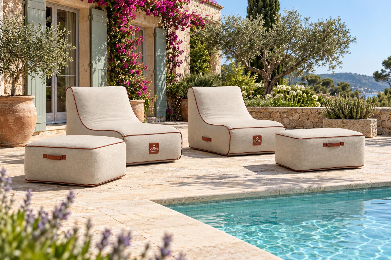 Fauteuils et poufs gonflables beiges disposés autour d'une piscine sur une terrasse ensoleillée devant une villa méditerranéenne avec végétation